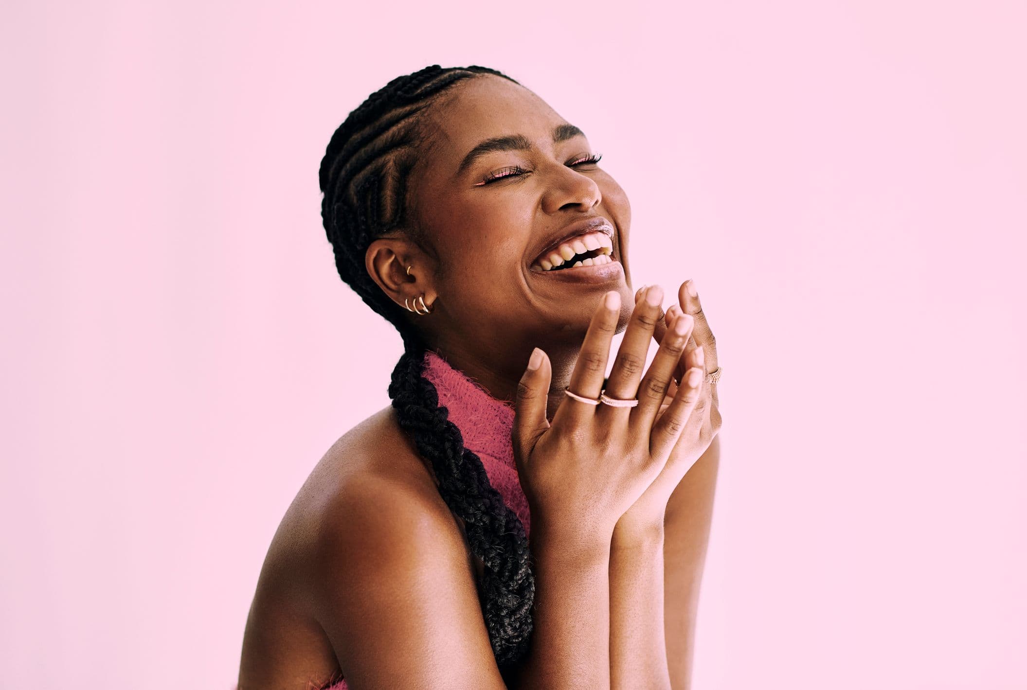 A Black woman with cornrows laughs joyfully with her eyes closed, hands clasped.