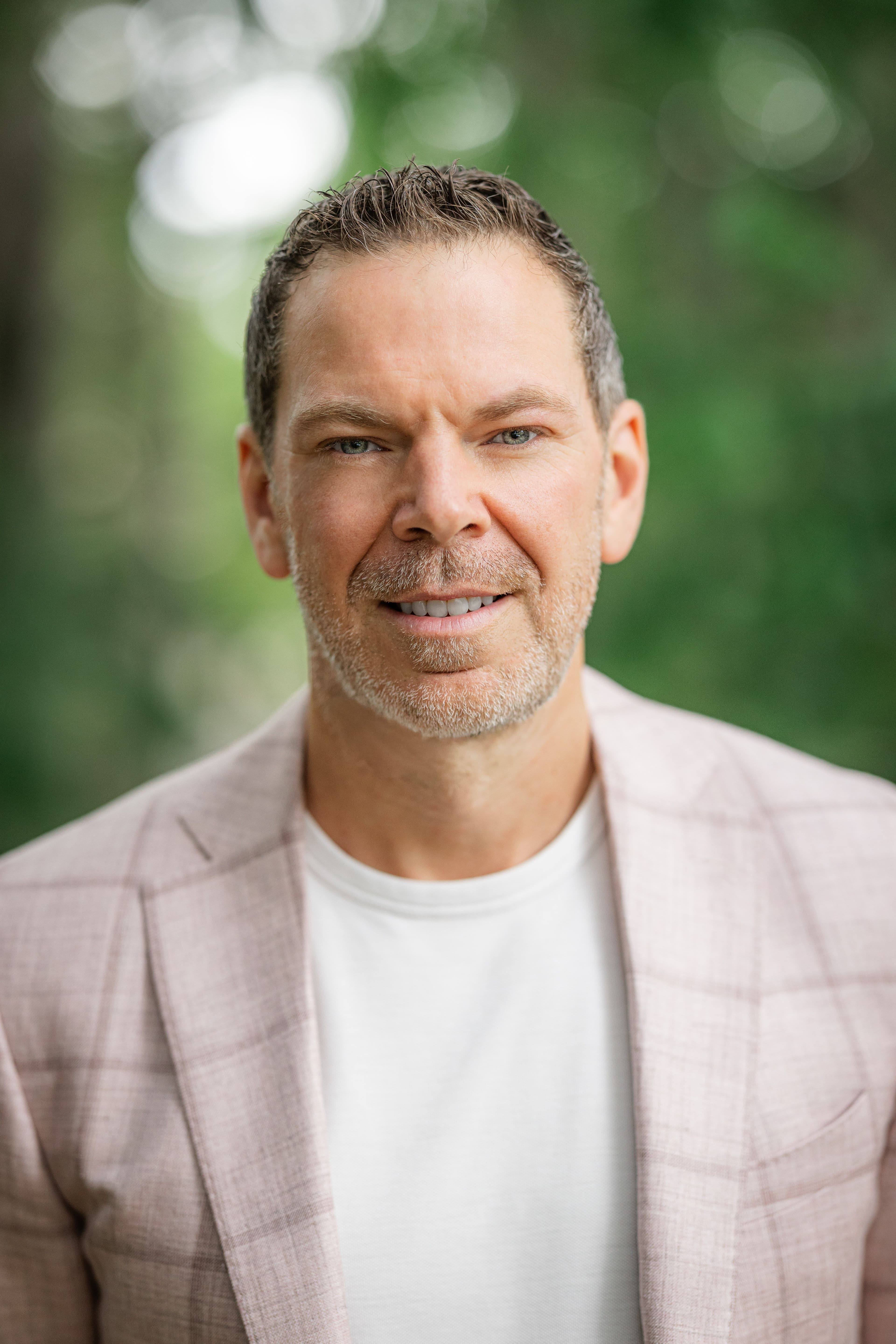 Headshot of a smiling man with short graying hair and light stubble, wearing a pink plaid blazer and white t-shirt against a blurred green background.
